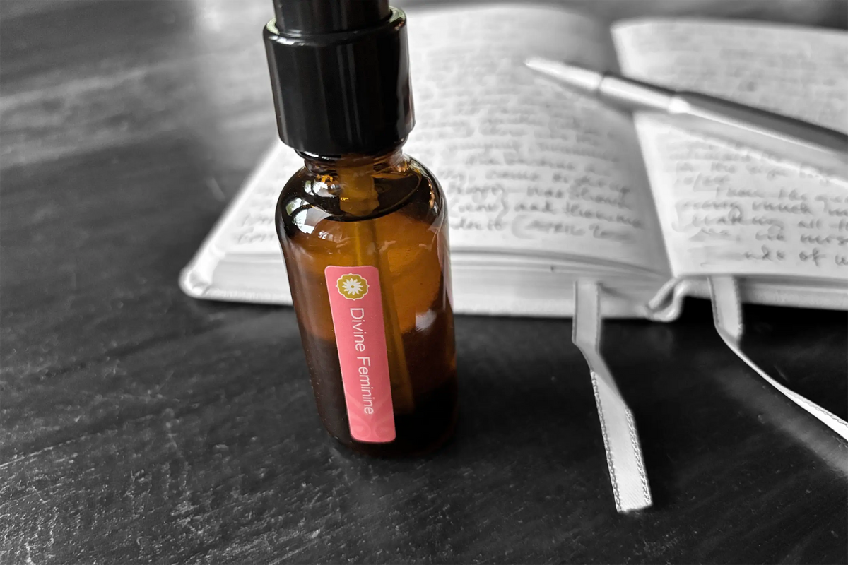 Amber glass bottle of BodyMantra Divine Feminine oil on a weathered wood surface with blurred image of a journal and pen in the background.