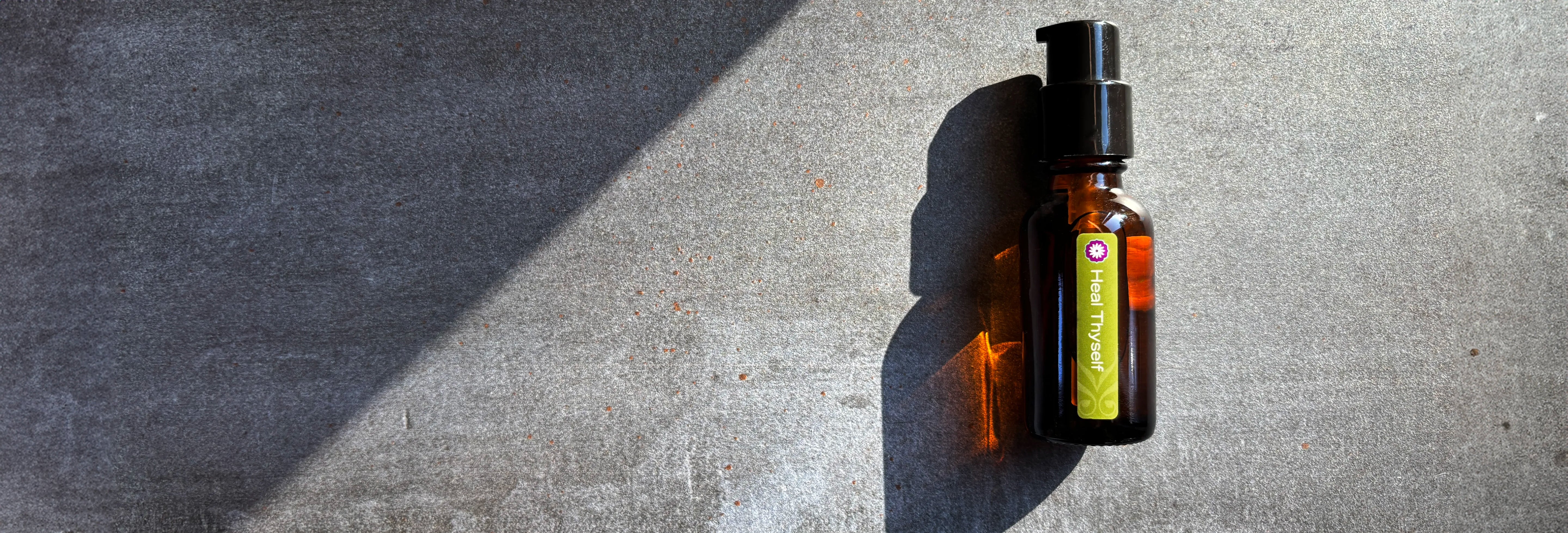 An amber glass bottle of BodyMantra Heal Thyself oil blend featuring refracted sunlight on a textured gray concrete surface with a strong diagonal shadow