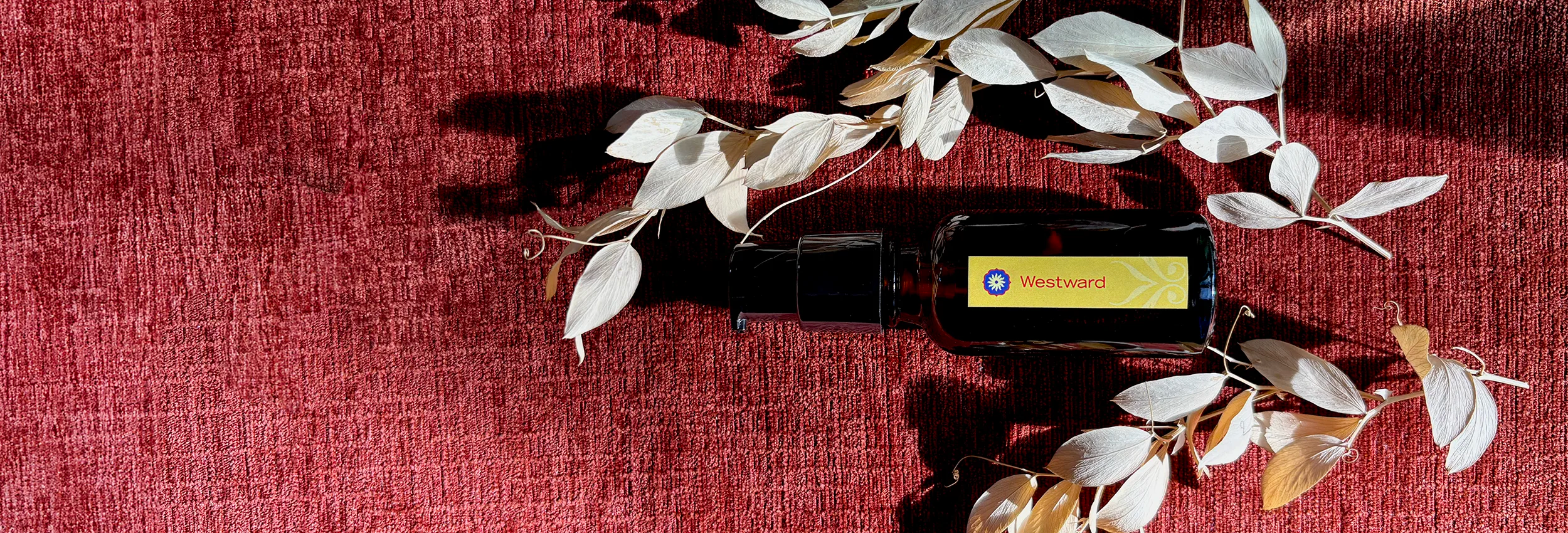 An amber glass bottle of BodyMantra Westward oil with a dark gold label on a textured rust-orange fabric background, surrounded by twigs with small dried ivory leaves.