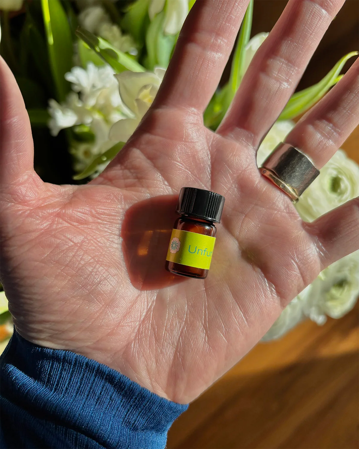 Hand holding a small vial of BodyMantra Unfurl oil blend with white spring flowers in the background.