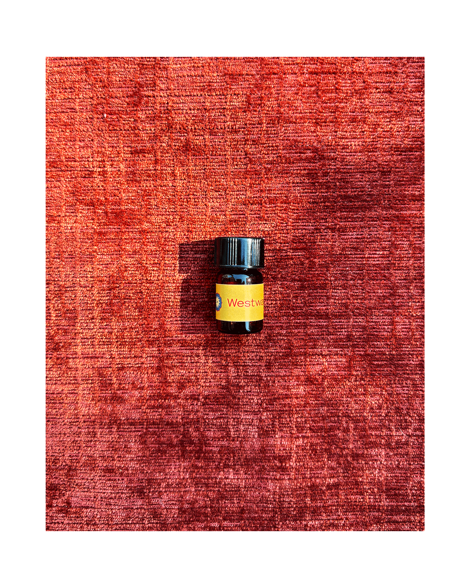A small amber glass vial of BodyMantra Westward oil with a dark gold label, placed on a textured rust-orange fabric background.