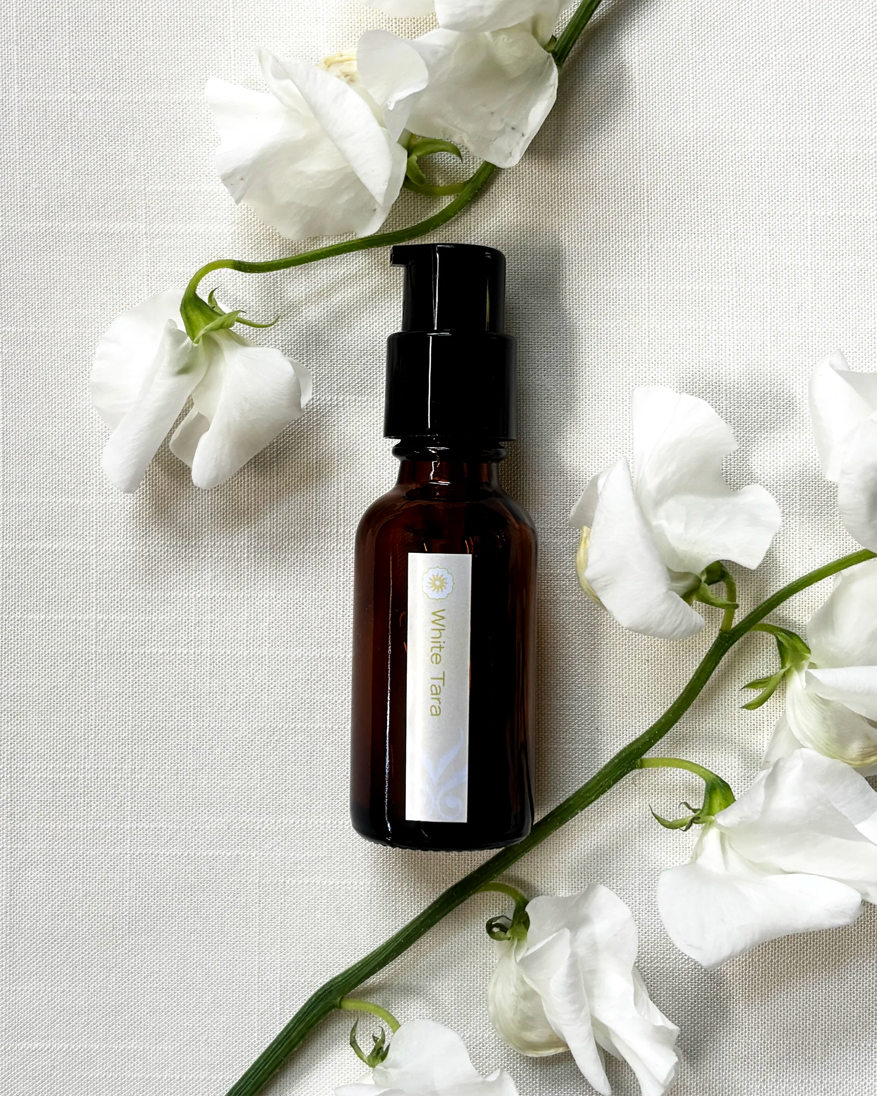 An amber glass bottle of White Tara oil by BodyMantra, featuring a minimalist label, set on soft-white linen, and surrounded by pure-white sweet pea flowers.