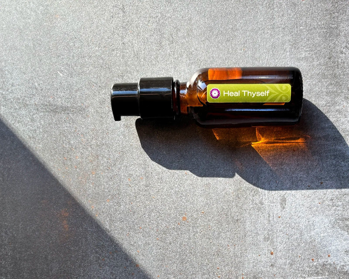 An amber glass bottle of BodyMantra Heal Thyself oil blend featuring refracted sunlight on a textured gray concrete surface with a strong diagonal shadow