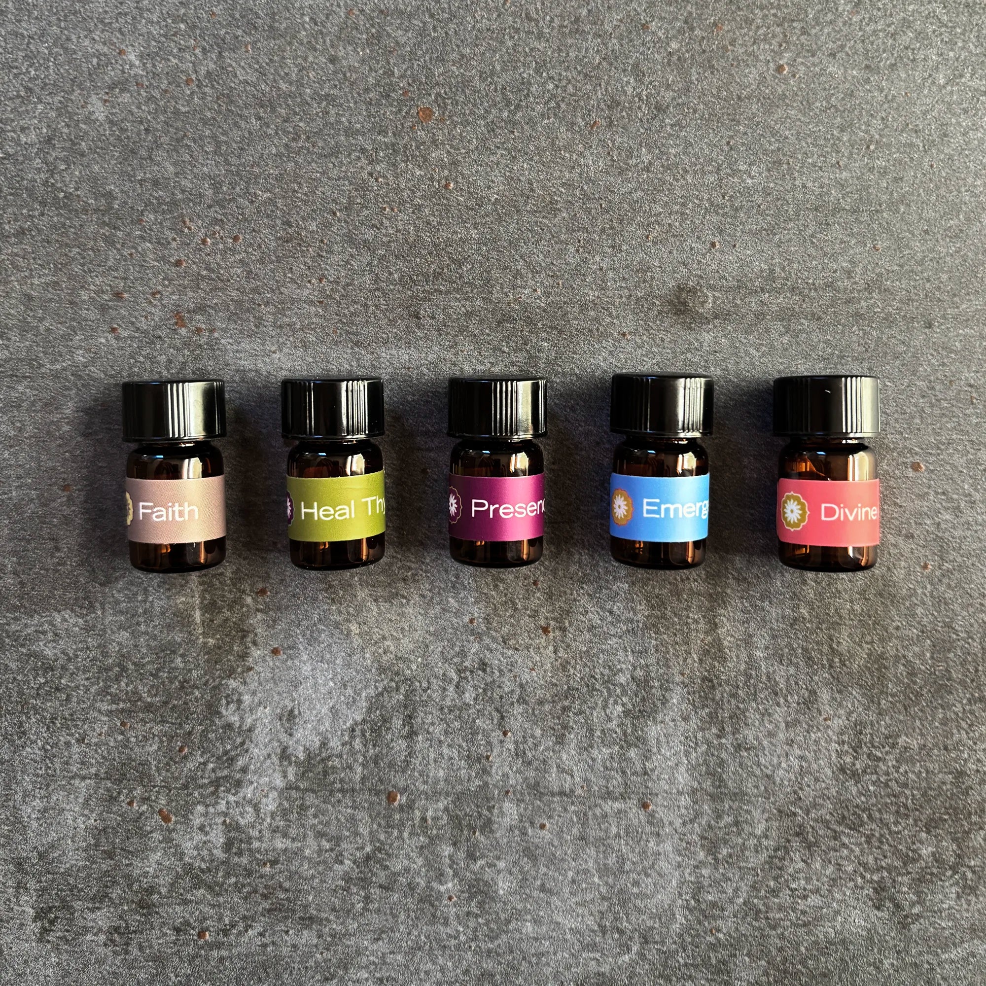 Five small amber vials lined up on a textured gray surface featuring labels of oil blends by BodyMantra: Faith, Heal Thyself, Presence, Emergence, and Divine Feminine.