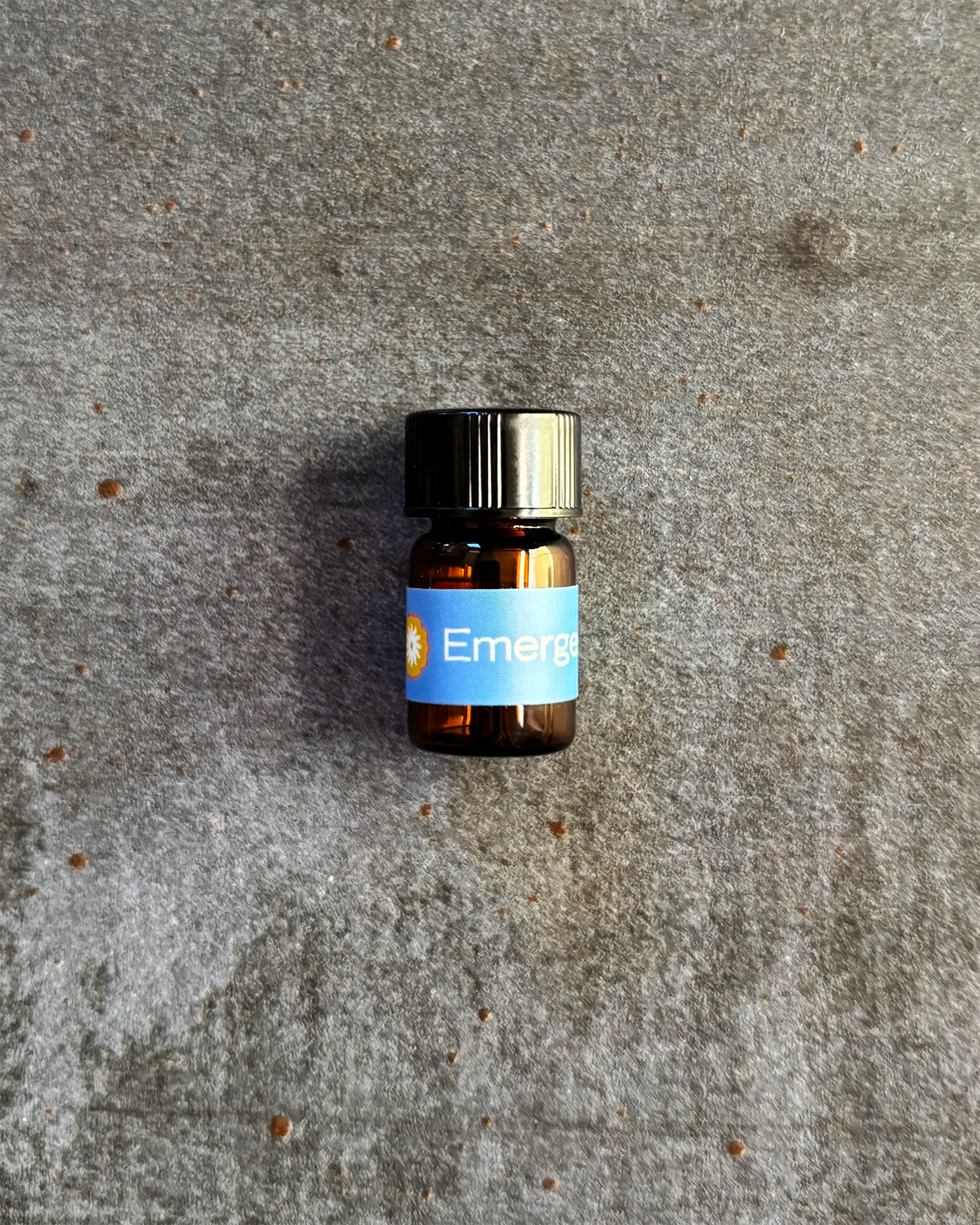 Small amber vial of BodyMantra Emergence oil blend on textured gray stone background.