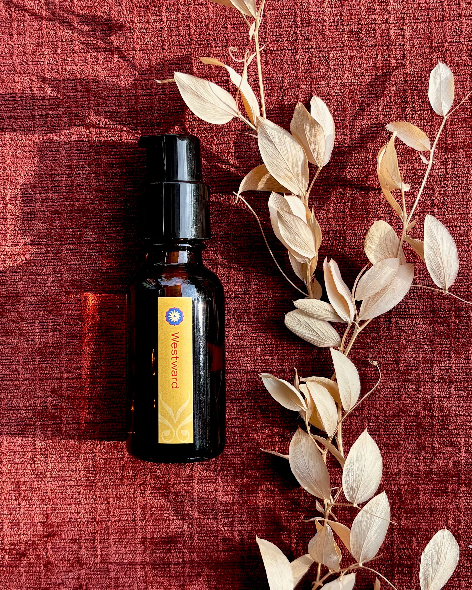 An amber glass bottle of BodyMantra Westward oil with a dark gold label on a textured rust-orange fabric background, surrounded by twigs with small dried ivory leaves.