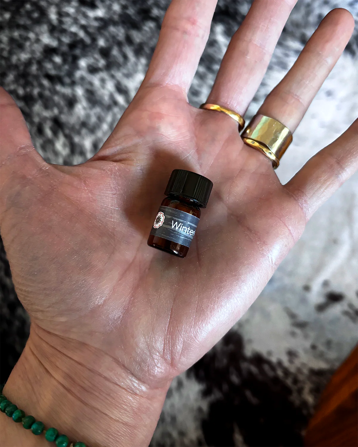 Hand holding a small vial of BodyMantra Wintering oil blend with a blurred black and white background pattern.