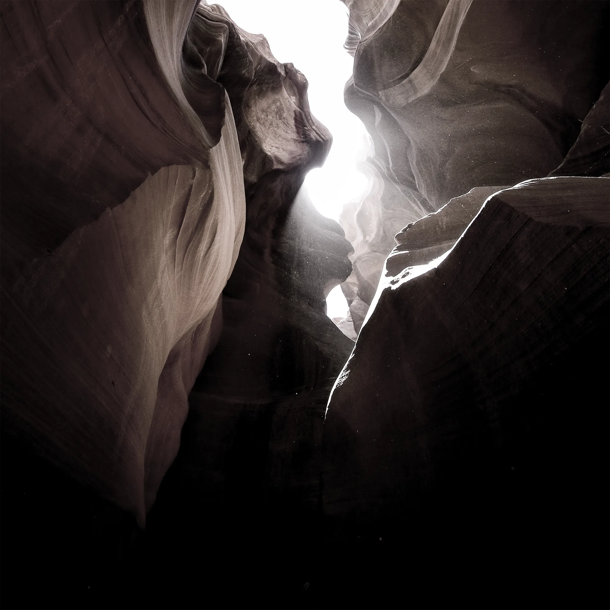 Photo angled looking up through a narrow canyon slot toward bright sky, warm sandstone walls curving inward.