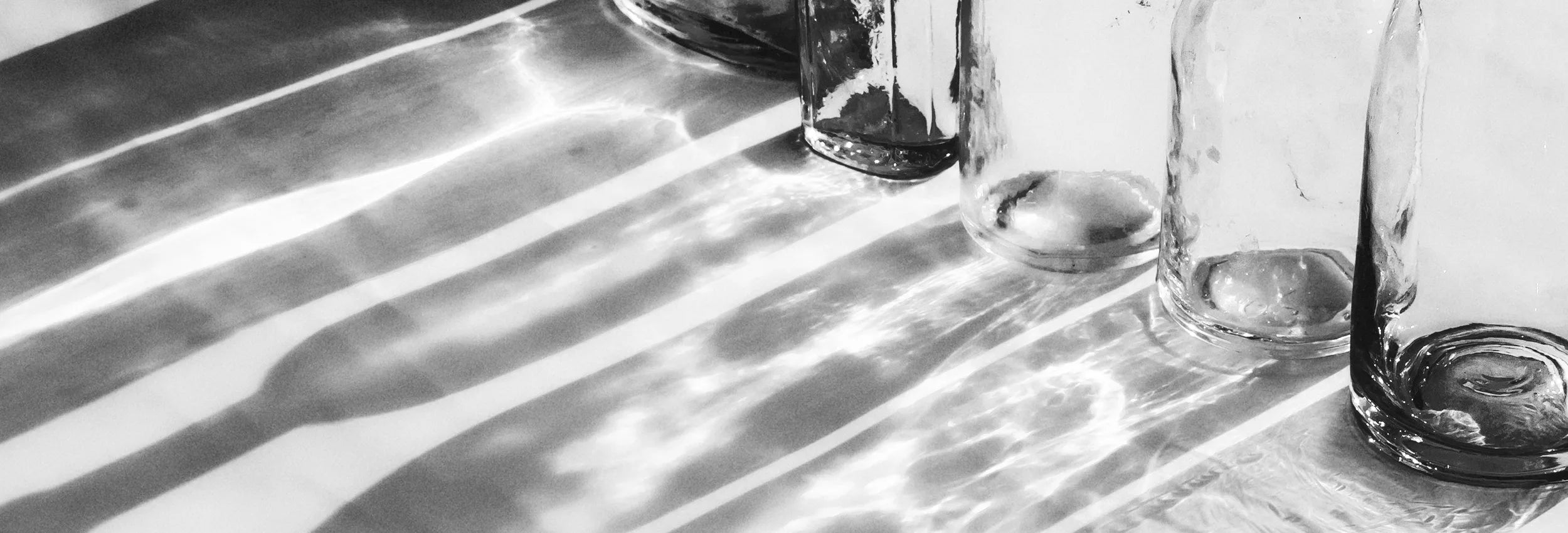 A black and white photograph of several clear glass bottles arranged on a reflective surface, casting long, bold shadows across a white background.
